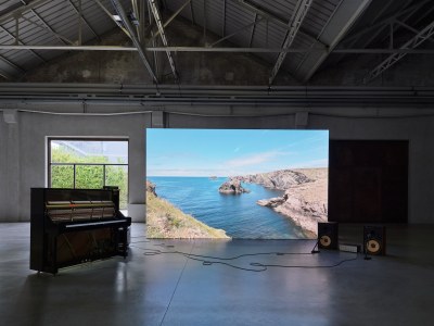 A piano is connected to two speakers on the floor, next to a large screen that shows the ocean from above.