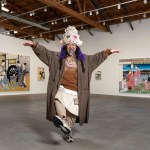 A bearded Japanese man in a robe, tall hat, raises his arms and looks at the camera in front of two paintings.