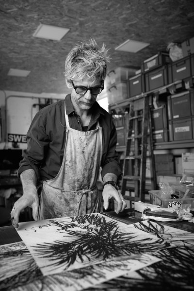 Black and white photo of Stephen Thomas Gallagher, wearing an apron and holding a charcoal stick. He looked down at some new drawings.