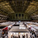 Aerial view of an art fair, with people crowding stalls. It's located in the Park Avenue Armory, which has a huge dome.