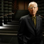 A middle-aged white man with white hair stands in a dark environment, wearing a suit and clear-framed glasses.