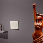 View from a museum exhibit showing detail of a wooden sculpture of a woman with one arm raised and the other crossed over her head to support the other. Following are two smaller sculpture studies and drawings of the work.