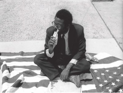 Archival photo of Pope.L sitting on an American flag and eating a newspaper.