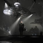 A person is walking in a future space, with a satellite-like object hanging overhead and spotlights pointed at different places.