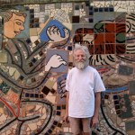 An old man with white hair and white beard, artist Isaiah Zagar, smiles in front of the mosaic artwork he created