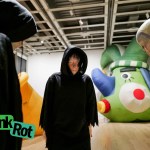 Three sculptures of hooded figures crowd in front of a colorful, enlarged sculpture of a court jester.