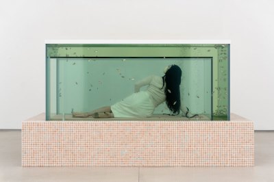 A woman poses in a fish tank with a cutout of a mattress. Fish swim back and forth in the water. 
