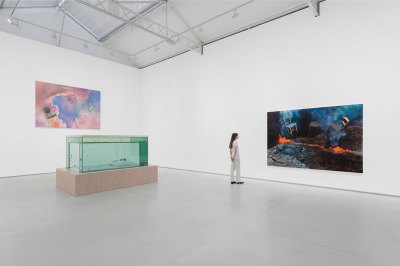 A woman looks at a painting of floating chairs in a volcanic landscape. There is a fish tank sculpture on a tile base behind her, and a similar painting of a chair on the wall. 