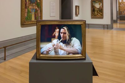 View of television in golden frame showing a man in white robe playing the flute. At the back is a museum gallery with early Renaissance gold leaf paintings. 