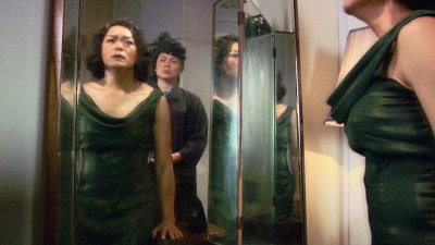 The film still shows a cross-dressing man in a green skirt looking into a mirror, with another person behind him. 