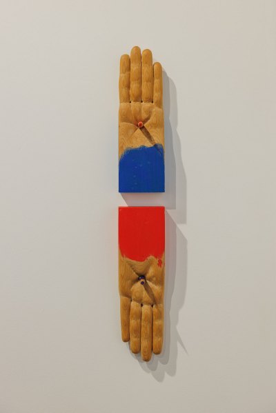 Wooden sculpture of two palms (without thumbs) with a stigmata-like thing in the middle. One is painted blue at the cuffs, the other is painted red at the cuffs. 