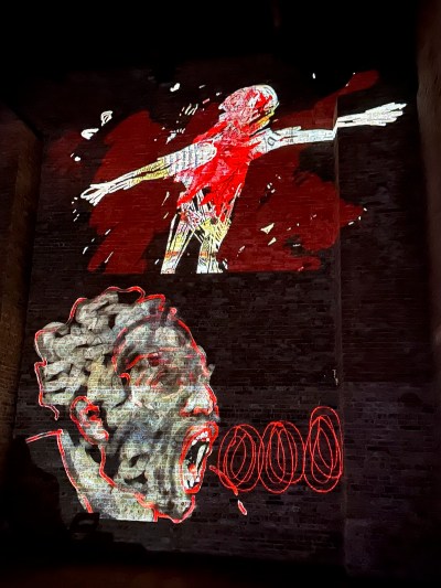 A man spreads his arms and a spitting face is projected onto the bricks.