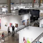 Photo shows rows of white-walled booths with artwork hanging on the walls and visitors in the aisles at the Expo Chicago Art Fair