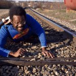 Pope.L wears a Superman suit and crawls across the railroad tracks.