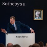 A man dynamically stands behind a podium and uses the word "Sotheby's" printed on it.