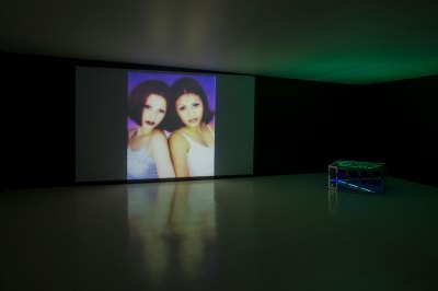 A projection of a mall photo from the '90s of two Latinas on a screen with a sculpture at right. 