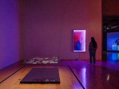A woman looks at a mirror work in the right background. At right a floating work with archival materials. 