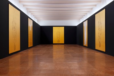 Gallery with paintings of tall upside down figures against golden background.