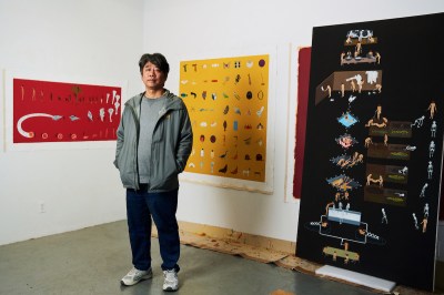 A man in a gray jacket stands next to a painting of a group of naked people performing various activities with milk.