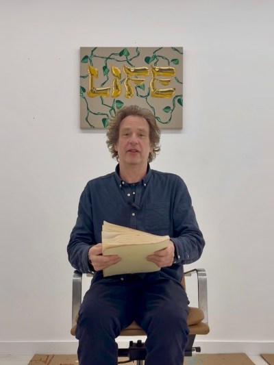 Joel Meisler sat in a chair with a bound script in his hands. Behind it is a word painting of the word