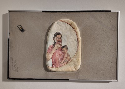 Framed mixed media piece on gray textured surface. A watercolor painting on an irregularly shaped piece of handmade paper in the center depicts a woman in a pink top holding a child and holding a mobile phone, as if taking a selfie in the mirror. There is a small SD memory card taped to the upper left of the composition.
