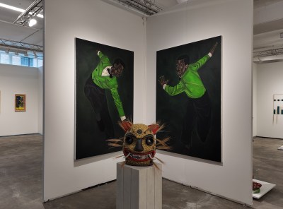 The gallery booth features two large paintings with dark backgrounds, each depicting a figure wearing a bright green jacket that captures dynamic, dance-like poses. In front of the painting, a sculptural mask on a gray base has a yellow texture on its surface, with large black eyes, teeth, red ears and radiating straw-like hair.