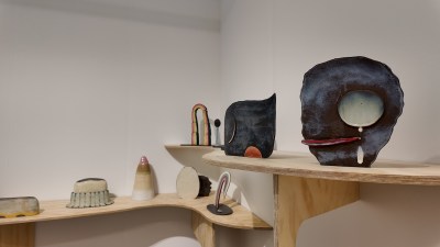 Small ceramic sculptures are displayed on curved plywood shelves mounted on the white gallery walls. The works range from dark-glazed abstract flat forms with circular cutouts and dripping details to smaller objects including iridescent arches, cones, and egg-shaped pieces in soft earth tones and pastels.