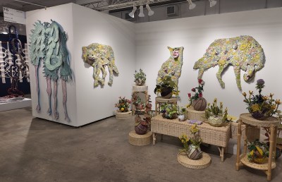 On the left wall is a large wall-mounted sculpture of a pale green fantasy creature with dangling legs. The booth on the right displays large animal silhouettes – resembling wolves or hyenas – made from collaged floral fabric and paper, mounted on a white wall. Wicker furniture and stands feature sculptural flowers growing from deflated sports balls.