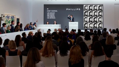 Phillips Auction House was auctioning Lot 15 (an Andy Warhol work) live, and the place was packed with people. The auctioneer stands on a podium with one arm raised, with a telephone bidder on his left. The price board displays data for multiple currencies (USD, EUR, GBP, CHF, HKD), and the screen to the right displays a portrait of Warhol—a grid of repeating portrait images in his signature style. Spectators sat in rows of white chairs, mostly watching from behind.