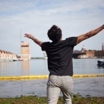 A man wearing a black T-shirt and plaid pants faces the lagoon, his arms stretched out at his sides and his head tilted toward the sky.