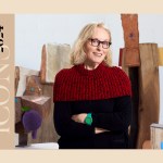 An older while woman with shoulder-length blonde hair, wearing a black and red sweater, has her arms crossed and is standing in a space with wood sculptures in the background