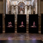 Five screens show a woman singing in a 12th century church below a mural.