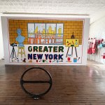 There is a bicycle rack in front of the mural, which features a bust, a camera, an easel and small paint images, with a view of the city through the windows.