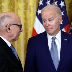 Two old men talk to each other in a room with yellow curtains and an American flag.