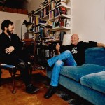Two white people sitting and talking, one on a chair and one on a couch, smiling, with bookshelves and artwork in the background.
