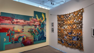 The gallery booth displays two large works side by side. To the left is a multi-panel painting in bright colors of teal, red and pink, depicting figures jumping or diving at the water's edge with beach balls and industrial crane hooks. To the right, a large textile wall in orange, gold and blue hangs with a grid pattern spread throughout, made of densely woven and knotted fibers. Small framed works hang between them.