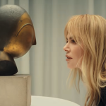 A woman with long blond hair and side bangs bends over a bronze head sculpture on a pedestal