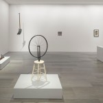 White walls of a gallery filled with ready-made works including a upside-down bicycle wheel on a stool and a shovel hanging from the ceiling.