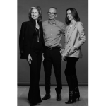 A black and white photo of a man wearing a gray shirt and dark trousers, flanked by two women. The woman on the left is wearing a dark suit and long chain. The woman on the right, in a light-colored double-breasted blazer and dark trousers.