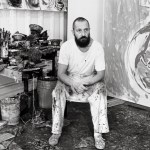 A bearded man sits in a painter's studio, wearing a white T-shirt and white, paint-stained pants.