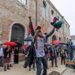 A group of people, some carrying umbrellas, outside a brick building. A man holds a Palestinian flag with his arms raised.