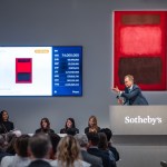 A man in a suit stood on a podium with a red abstract painting in front of him.
